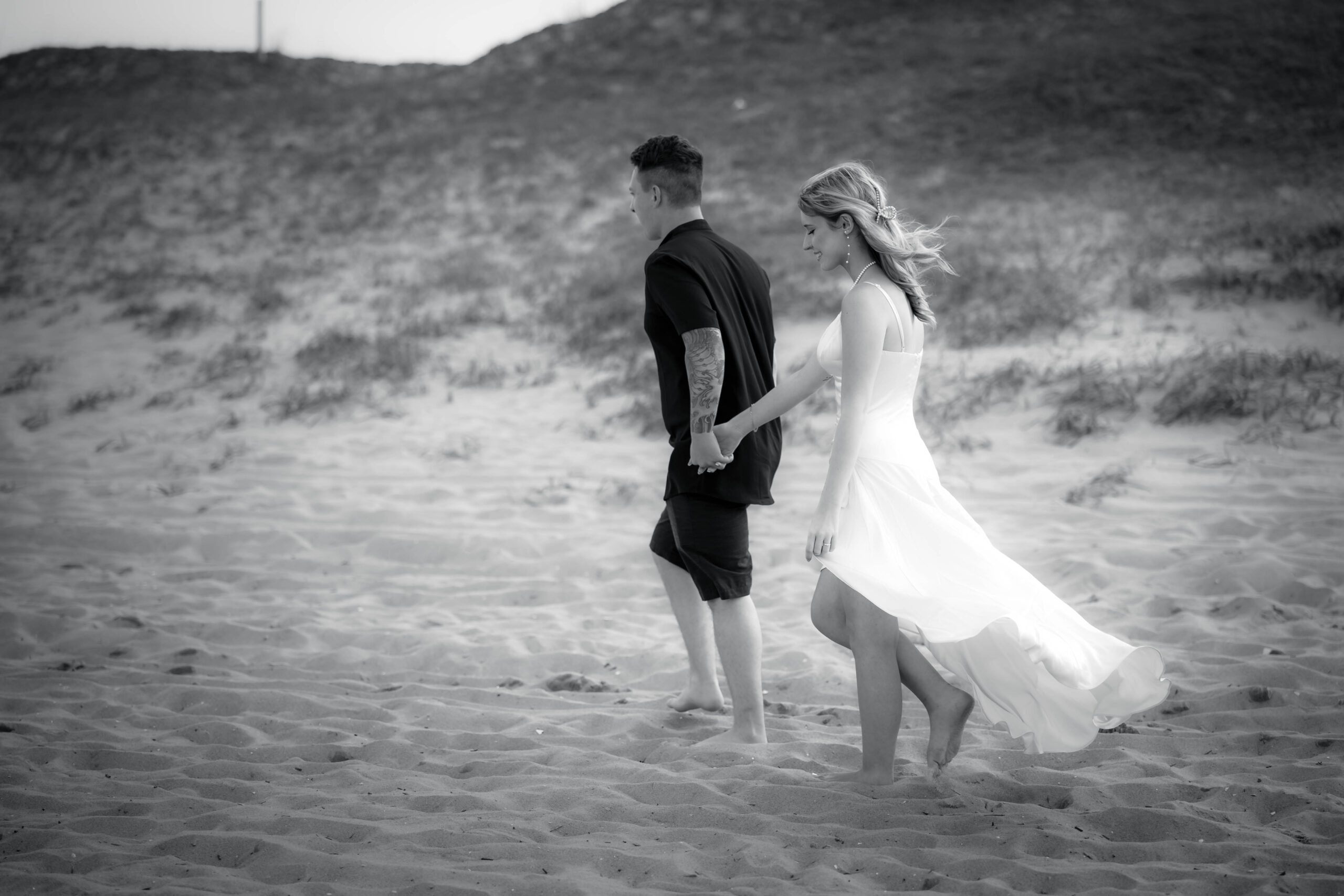 Black and white photo of a wedding day Bride and Groom. Bride dressed in a white gown flowing in the breeze of the ocean. Groom dressed in casual black. Bride and Groom walking hand in hand on the beach near the dunes.