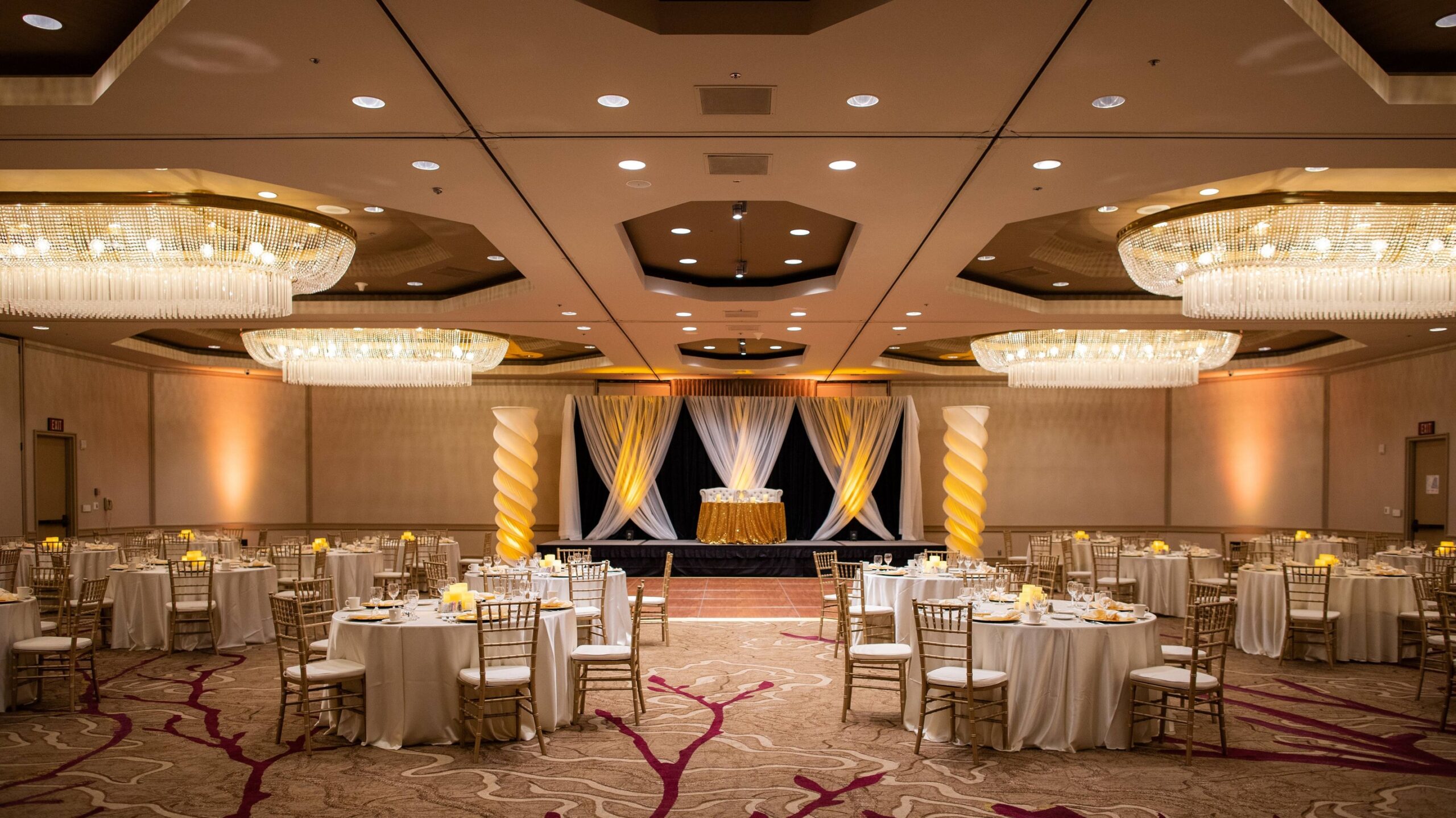 Large ballroom draped in warm lighting. Tables covered with white lines and adorned with yellow candles and yellow place setting chargers.