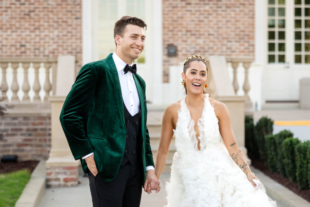 Bride and Groom facing camera but looking off to the side.  Bride wearing a full tulle drop v neck white wedding gown.  Groom wearing a green velvet tuxedo with white shirt and black bow tie.  Bride and Groom are holding hands and smiling and laughing.
