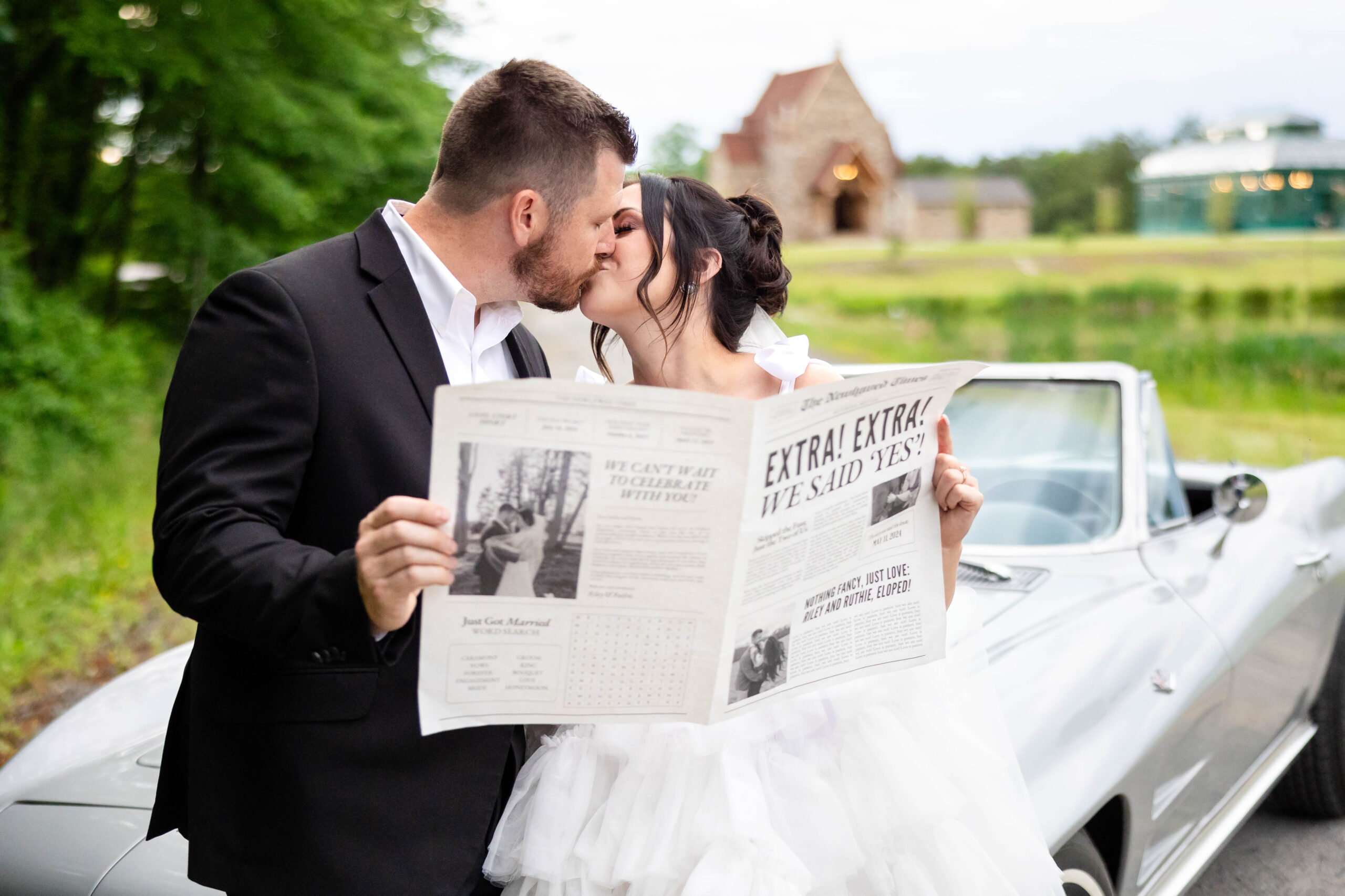 Bride and Groom with their classic convertible car. Bride dressed in a poufy short white wedding dress with ribbon shoulder ties. Groom in a black tuxedo. Bride and Groom are leaning on the front of the car and kissing. Bride and Groom are holding a newspaper with them as the headlines about just getting married.