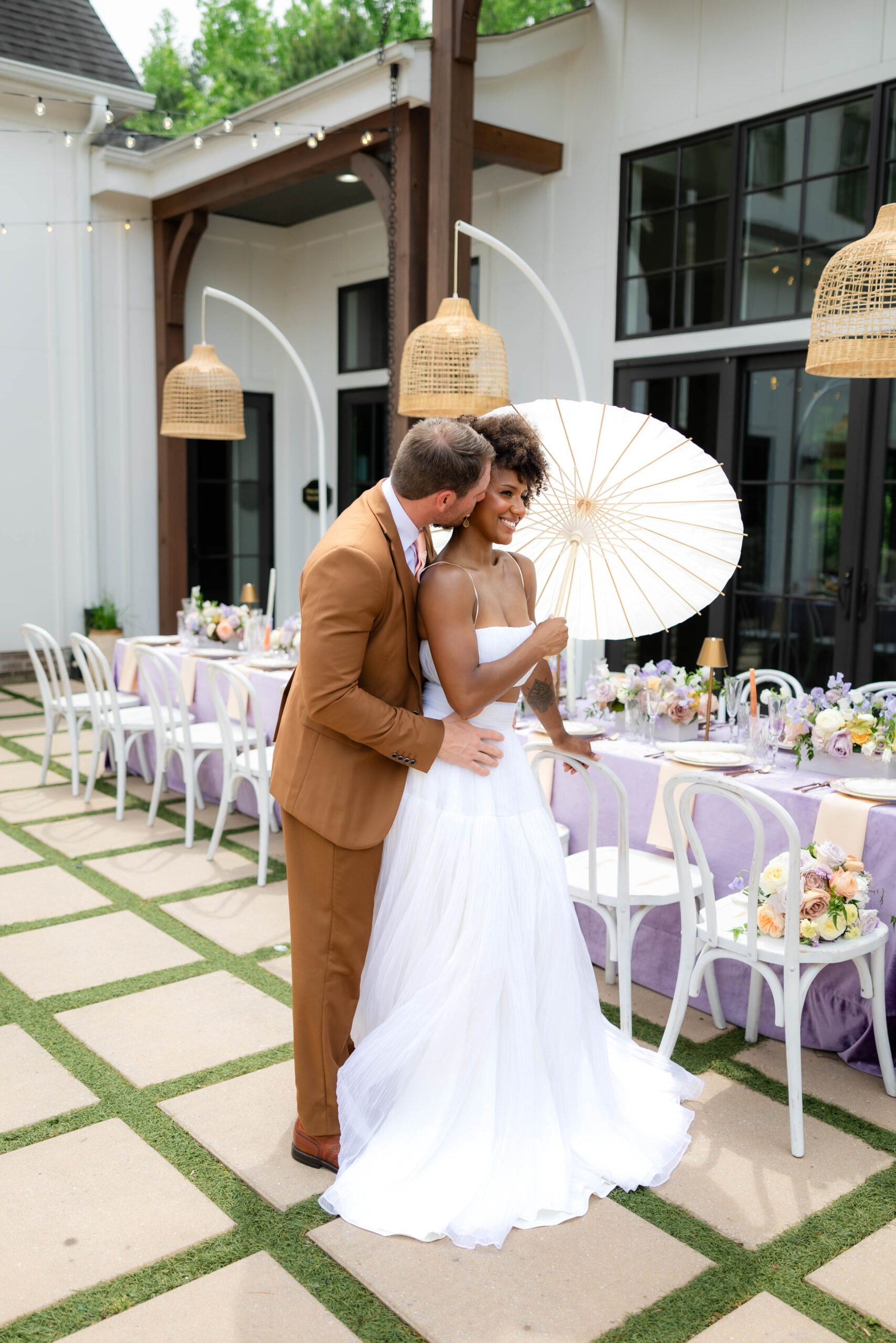 Bride and groom standing next to reception table. Bride holding a parasol. Groom leaning in to kiss the Bride's neck.