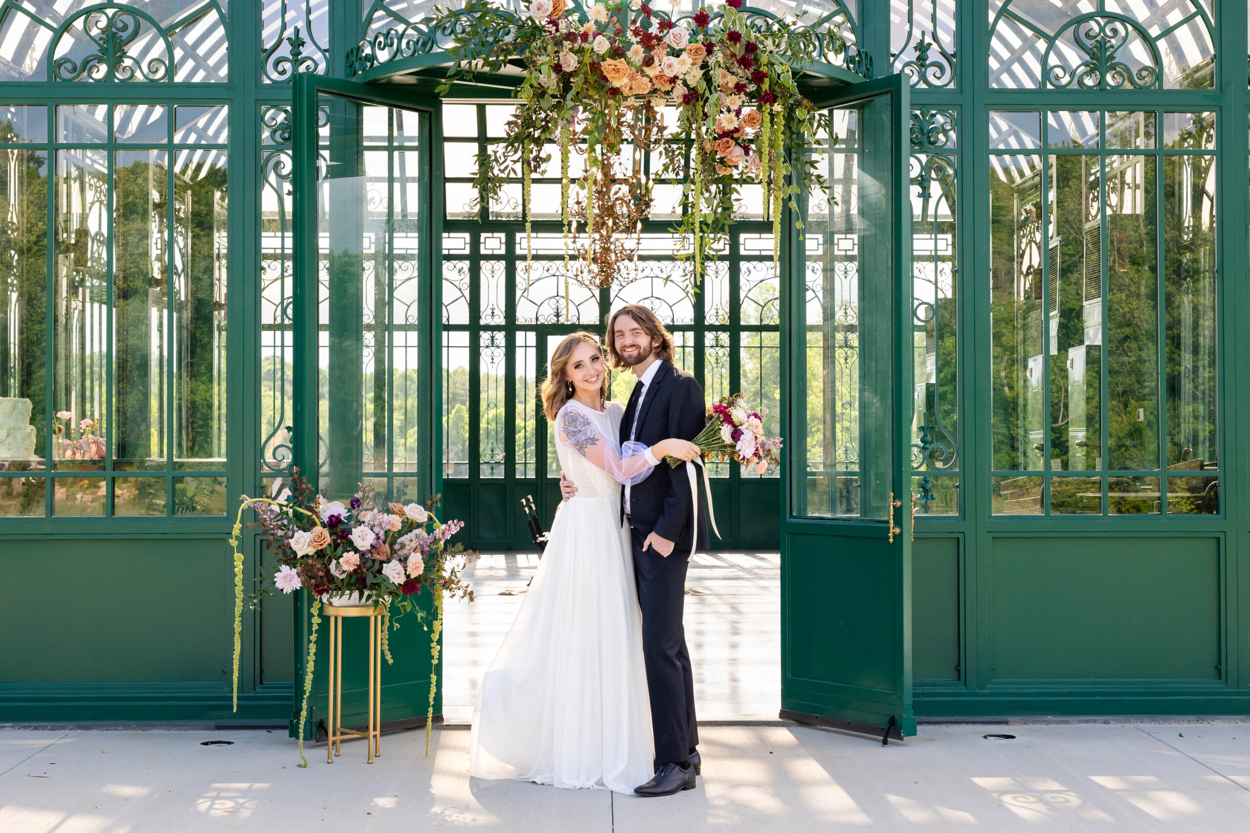 Bride and groom with florals framing the green atrium where the wedding reception was held. Bride and groom in a belly button to belly button embrace. Both facing the camera and smiling.