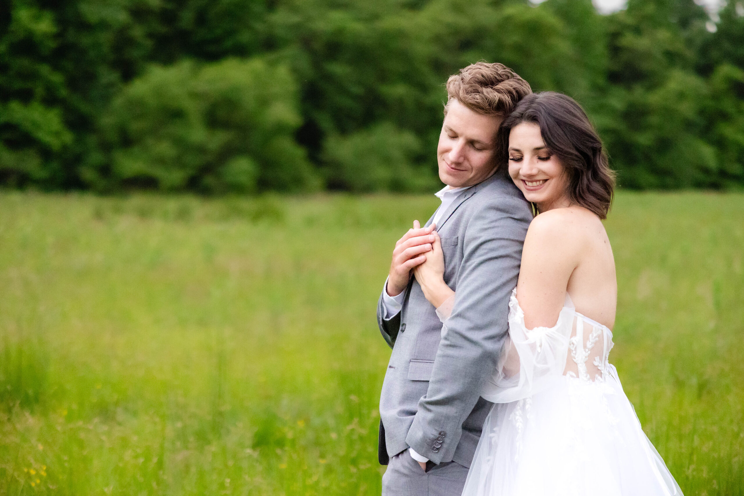 Bride and Groom in a field of spring grass. Bride is hugging her groom from behind as he turns and leans in to her. Bride in a custom strapless gown and groom in a light gray tuxedo. Photographed by Virginia Beach Wedding Photographer, Angela Foushee Photography