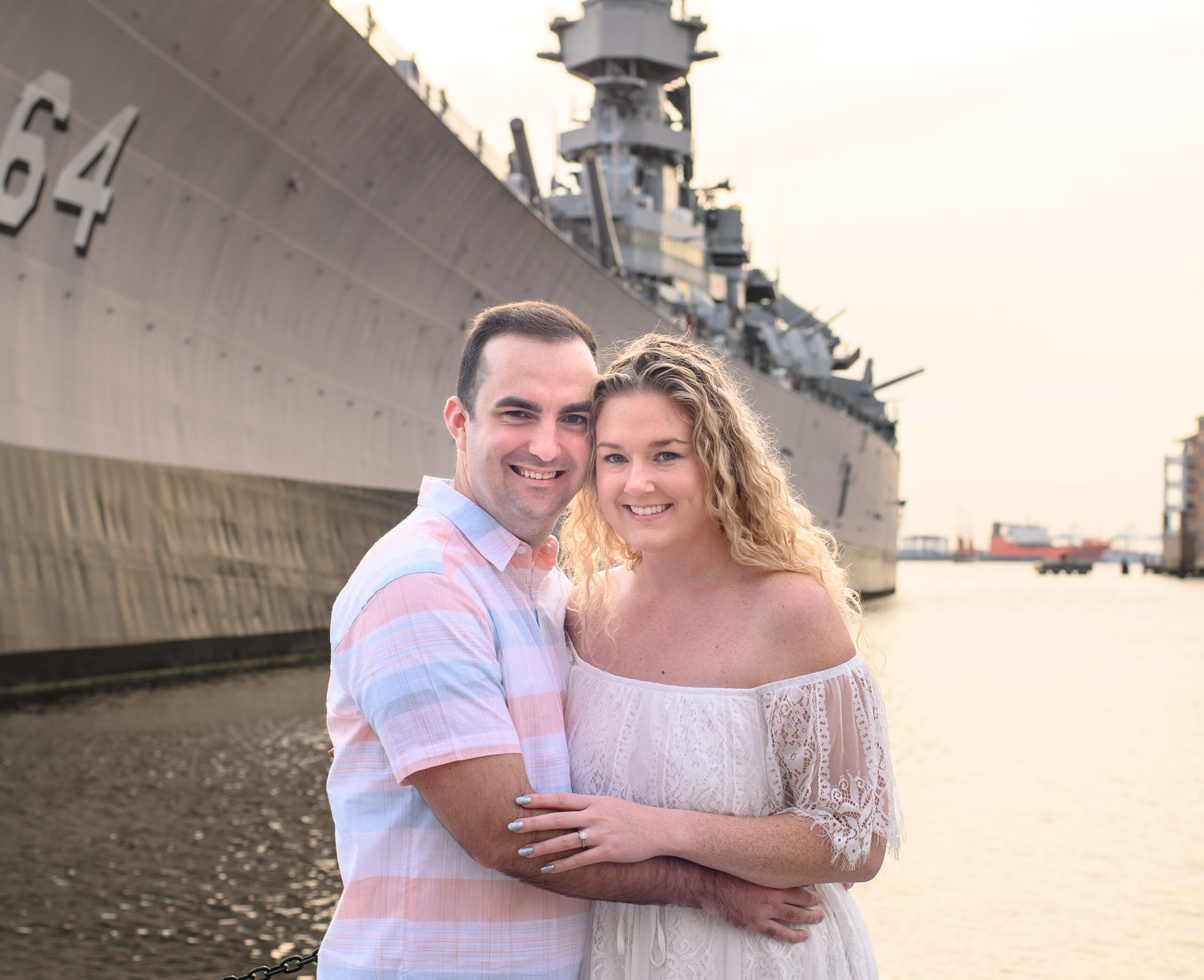 Newly engaged couple in a side embrace while looking at the camera and softly smiling. In the background is the battleship Wisconsin in Norfolk Virginia