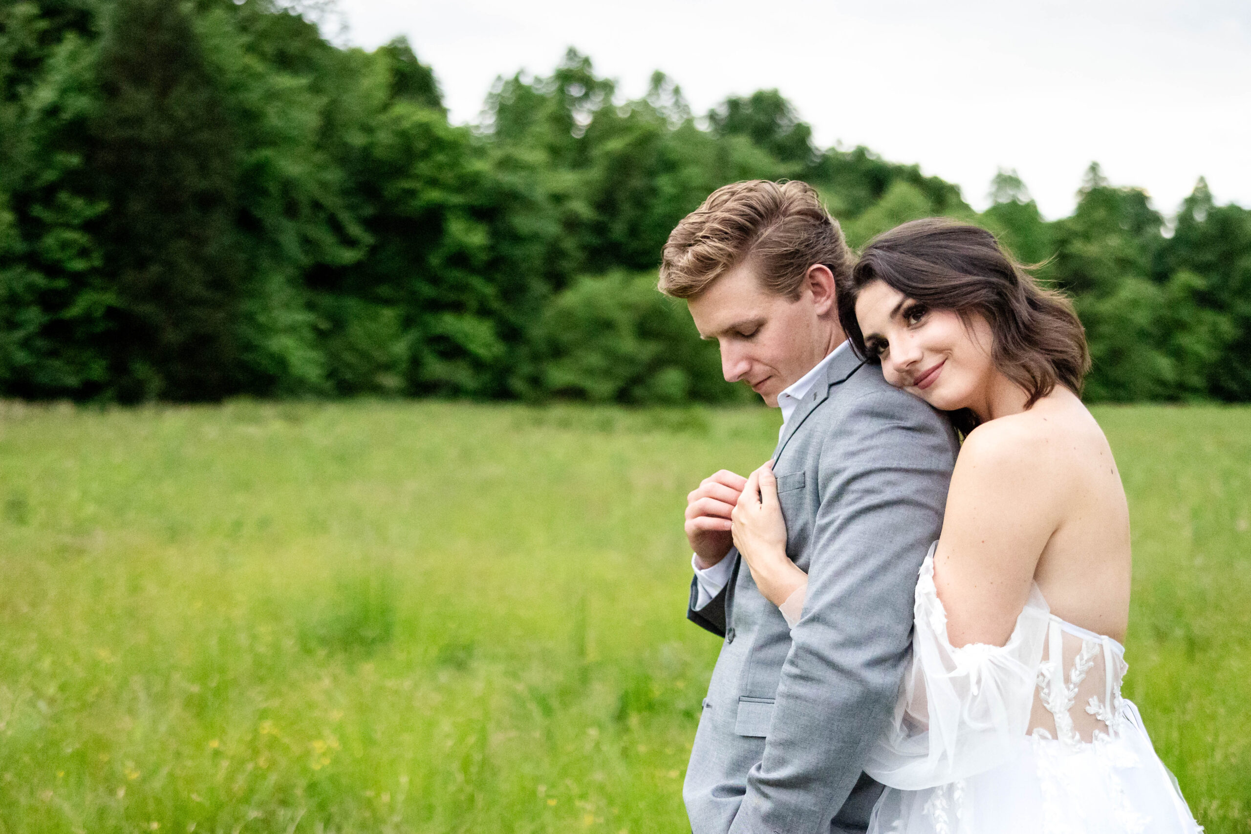 Bride and Groom in a field of bright summer green grass lined with trees in the distance. Bride is hugging her groom from behind as he gently touches her hand that is placed on his chest.