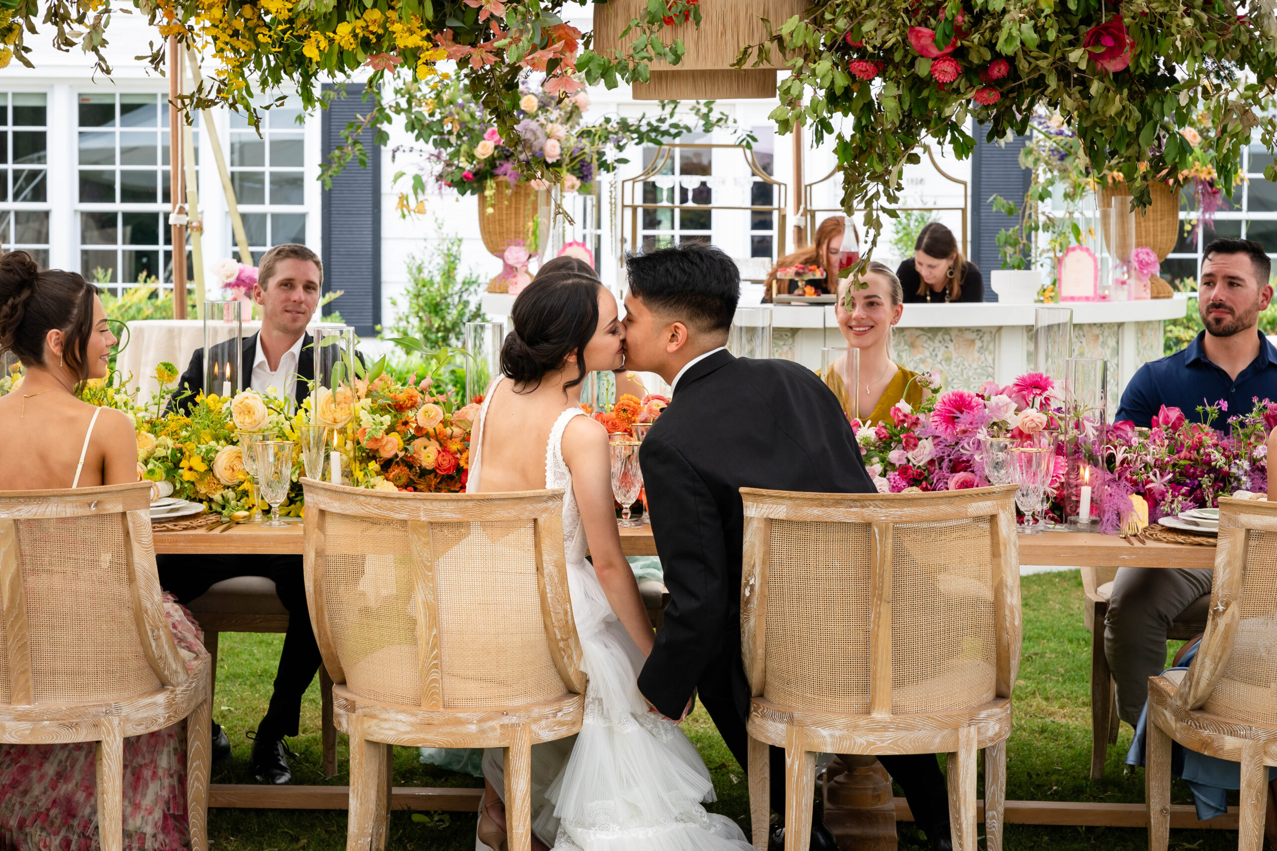 Wedding couple seated at sweatheart table that is draped with florals. Couple is seated at an outdoor tented reception. They are leaning in for a kiss as their wedding party looks on.