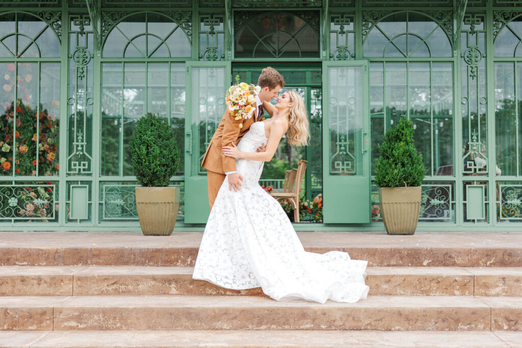 Bride and Groom in front of an atrium for formal portraits.  Bride in strapless gown and groom in light tan suit.  The groom is dipping the bride as he goes in for a kiss.  Bride is smiling up at her groom.  Photographed by a Virginia Wedding Photographer