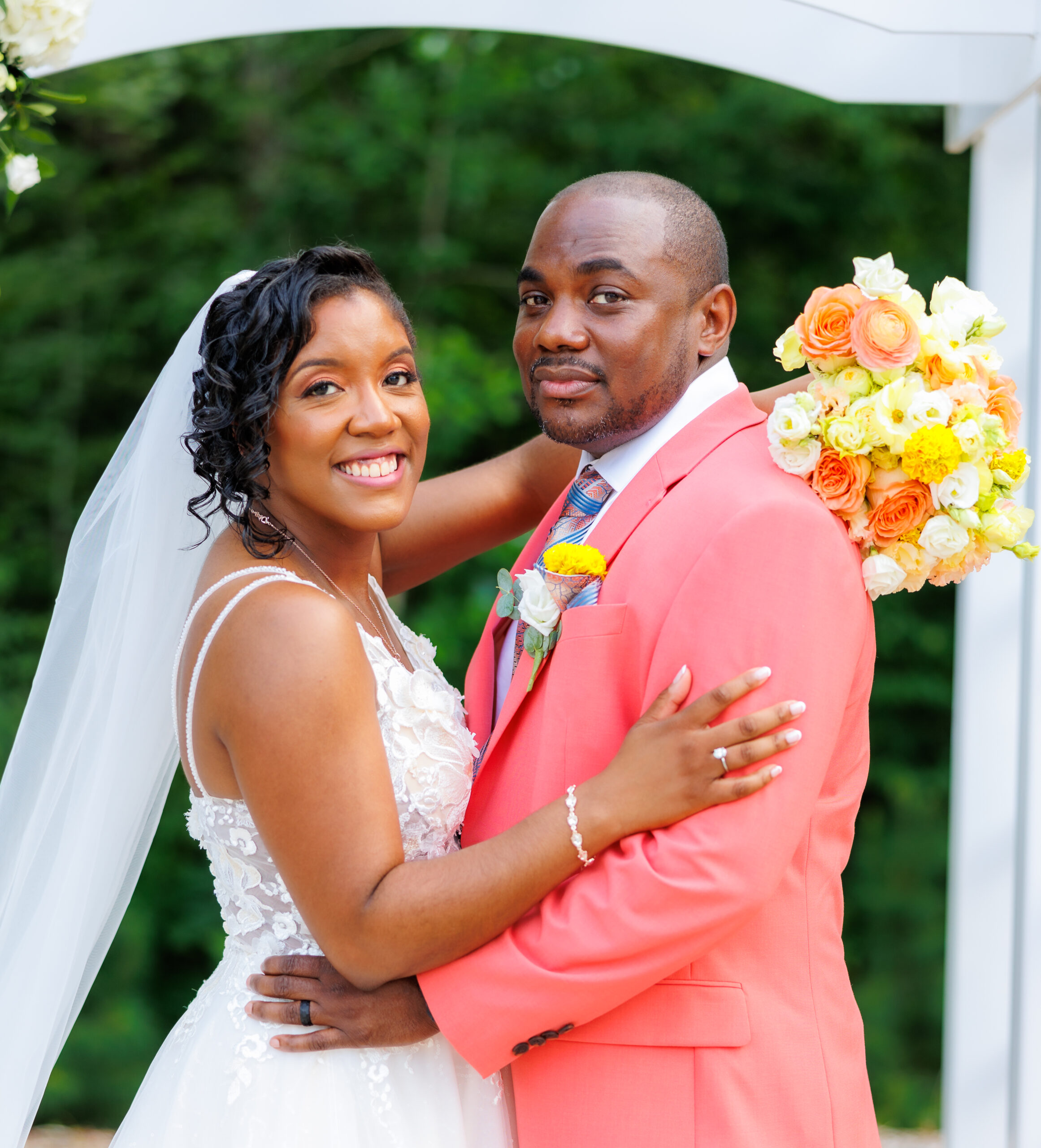 A bride and groom stand facing each other, smiling warmly at the camera. The bride wears a flowing white gown with peach accents and delicate lace details, and her hair is styled in an elegant waves updo adorned with pearls. The groom is dressed in a soft salmon-hued suit. They are surrounded by an arrangement of soft citrus florals, including pale oranges, yellows, and whites, with greenery accentuating the bouquet and decor. The background is softly blurred, emphasizing the couple and the vibrant, fresh flowers that add a light and airy feel to the photograph.