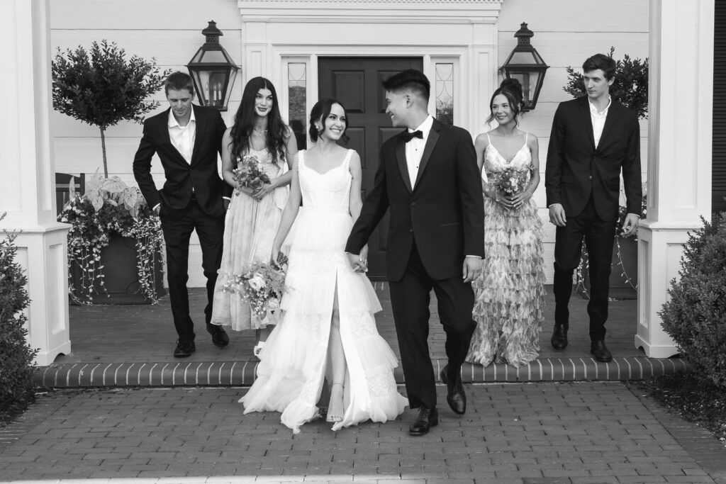 Bride and Groom and their wedding party outside of the estate venue.  Photo is in black and white.  Bride and Groom are holding hands walking out of the venue while their wedding party is following them closely behind.  