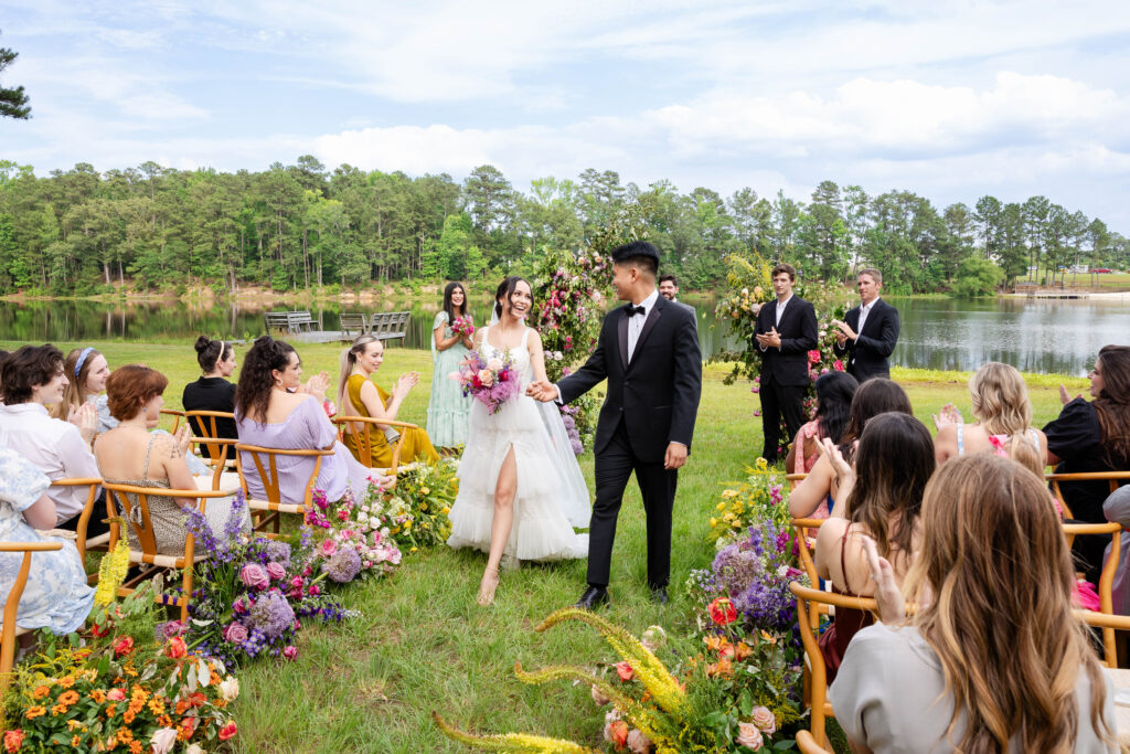 Bride and Groom exiting their lake front setting wedding ceremony.  The aisle is full of bright and colorful florals.  Bride is laughing at Groom as he takes her hand and begins to guide her back down the aisle to exit.  Taken by a Virginia Wedding Photographer
