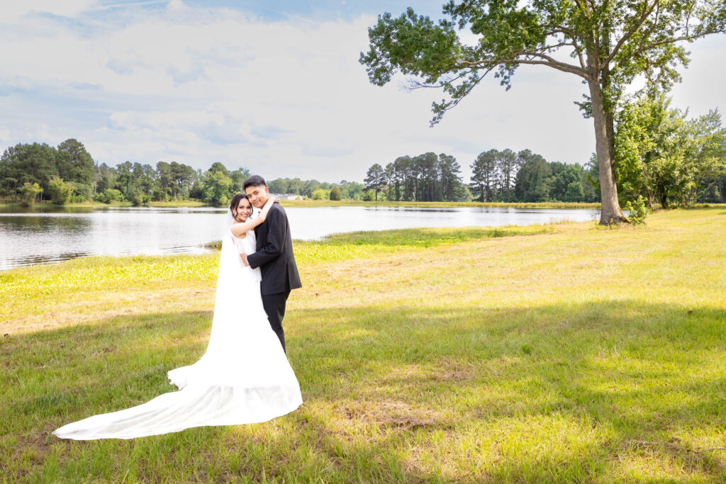 A bride and groom embrace on a sunny day by a serene lake, surrounded by lush greenery and trees, capturing a romantic moment in a picturesque outdoor setting