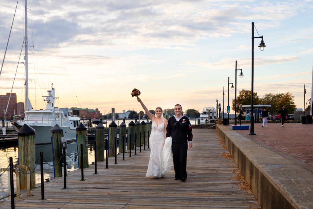 A joyful bride in a lace wedding dress and a groom in a navy uniform walk hand-in-hand along a wooden dock at sunset, with boats and a calm waterfront in the background. The bride raises her bouquet triumphantly, celebrating their special day.