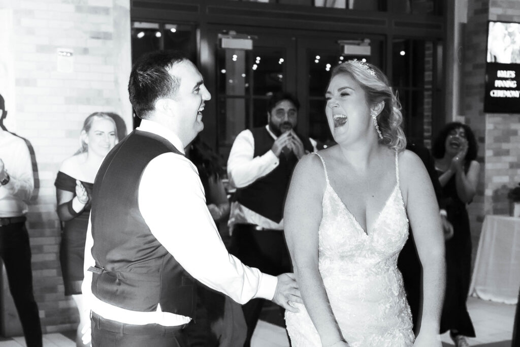 A joyful bride and groom share a candid moment of laughter on the dance floor during their wedding reception, surrounded by cheering guests in a black and white photograph.