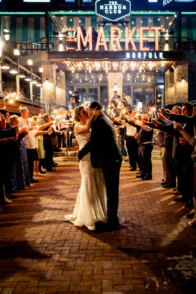 A bride in a sparkling white gown and a groom in a dark suit share a romantic kiss during their grand sparkler exit, surrounded by cheering guests holding sparklers. The scene is set in front of "The Market Norfolk" with warm lights and a festive atmosphere.