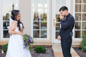 A bride in a white gown holding a bouquet of purple flowers smiles warmly as the groom, dressed in a black suit, wipes away tears during their emotional first look outside a building with large glass doors.