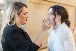 A makeup artist applies lipstick to a bride wearing a pearl-embellished wedding gown and elegant drop earrings, preparing her for the big day.