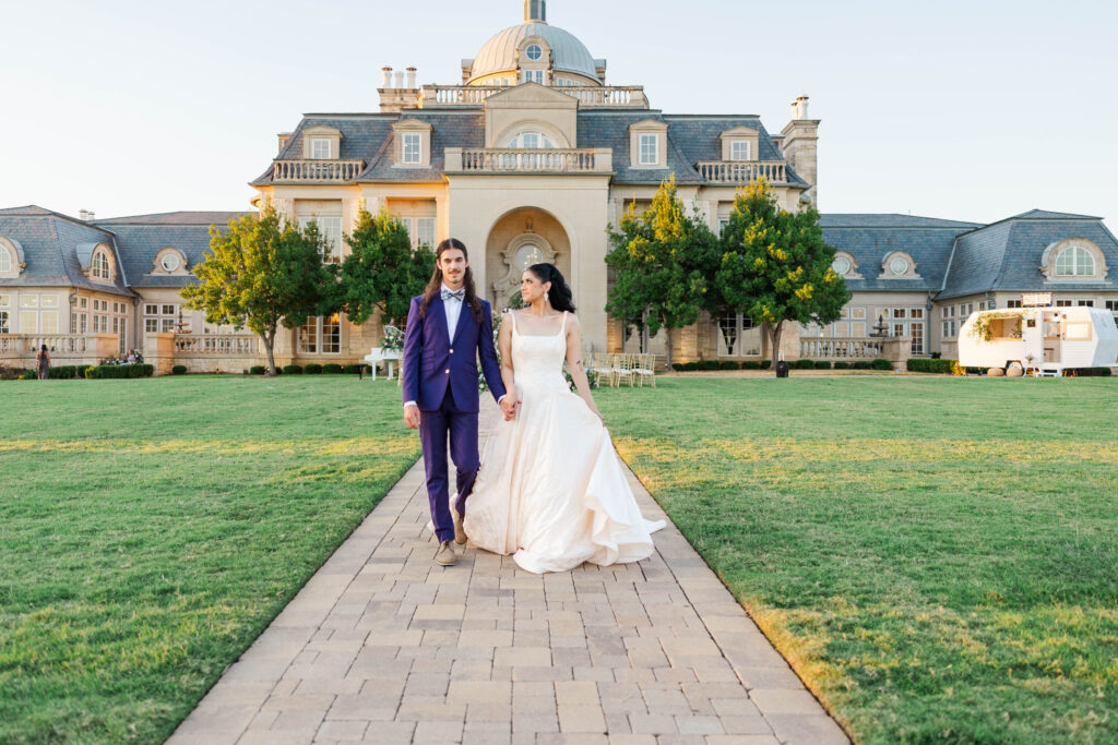 A bride in a flowing white wedding dress and a groom in a sharp blue suit walk hand-in-hand down a stone pathway in front of a grand estate with lush green lawns and elegant architecture.