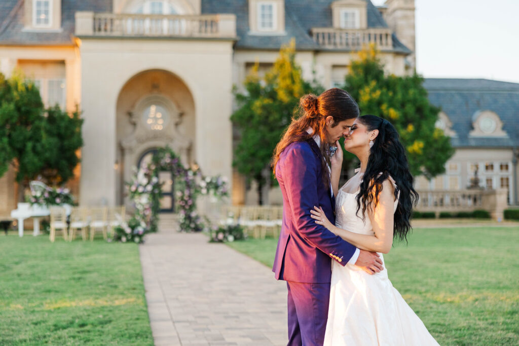 A bride in a flowing white gown and a groom in a deep purple suit share an intimate moment, gently touching foreheads and smiling, standing on a stone pathway in front of a grand estate with a floral arch and elegant outdoor seating in the background.