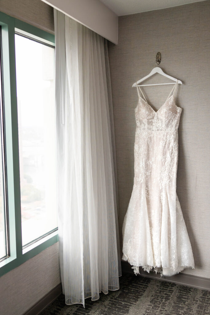 A Virginia Beach Wedding Photographer captures a stunning lace wedding dress with delicate floral details hangs on a white hanger against a neutral wall, next to a window with sheer curtains. Soft natural light highlights the intricate texture and elegance of the gown.