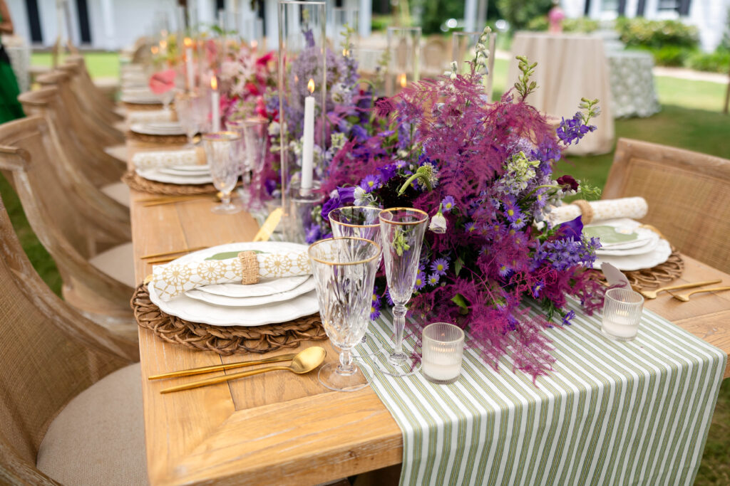 A beautifully decorated wedding reception table featuring a vibrant centerpiece of purple and pink flowers, gold flatware, crystal glasses with gold rims, and white plates with patterned napkins, set on a wooden table with a green and white striped table runner in an outdoor garden setting.