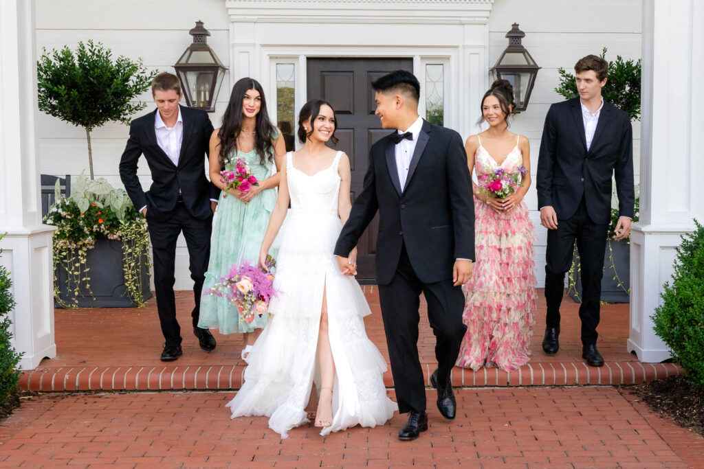 A bride in a tiered white gown and a groom in a black tuxedo walk hand-in-hand, smiling at each other, while their bridal party, dressed in pastel floral dresses and black suits, follows behind them on a brick pathway in front of a white building with black doors and lanterns.