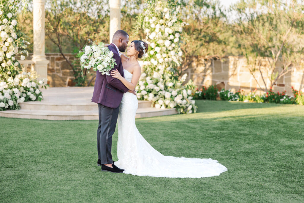 A bride in a fitted lace wedding gown and a groom in a burgundy suit embrace lovingly on a lush green lawn, surrounded by elegant white floral arrangements and a stone gazebo in the background, bathed in soft sunlight.