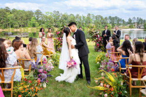 A bride and groom share their first kiss as a married couple during an outdoor wedding ceremony, surrounded by vibrant floral arrangements, seated guests, and a scenic lake backdrop under a bright blue sky.