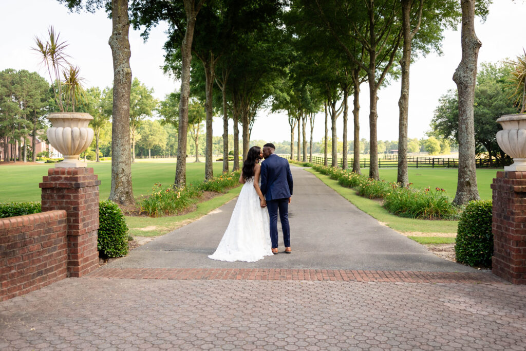 A bride in a flowing white gown and a groom in a navy suit share a romantic kiss on a tree-lined pathway, surrounded by lush greenery and elegant brick accents, creating a timeless wedding moment.