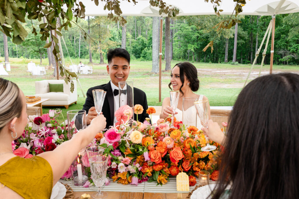 A joyful wedding reception scene with guests seated at a beautifully decorated table adorned with vibrant pink, orange, and purple floral arrangements. The bride in a white gown and the groom in a black tuxedo smile as they raise their glasses for a toast under a canopy of greenery and hanging flowers, with a white estate house in the background.