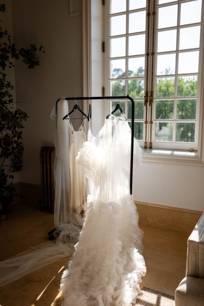 A delicate wedding dress with layers of soft tulle and ruffles hangs on a black rack near a sunlit window, accompanied by a veil and slip dress. The natural light highlights the intricate details, creating a romantic and elegant atmosphere.