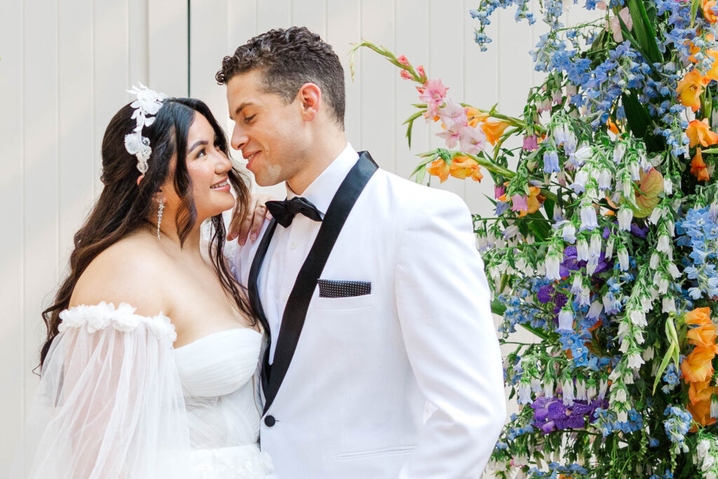 A bride and groom share a loving gaze on their wedding day. The bride wears an off-the-shoulder white gown with sheer sleeves and a floral headpiece, while the groom is dressed in a white tuxedo with a black bow tie. They are standing beside a vibrant floral arrangement featuring blue, orange, and pink flowers. Captured by wedding photographers in Virginia