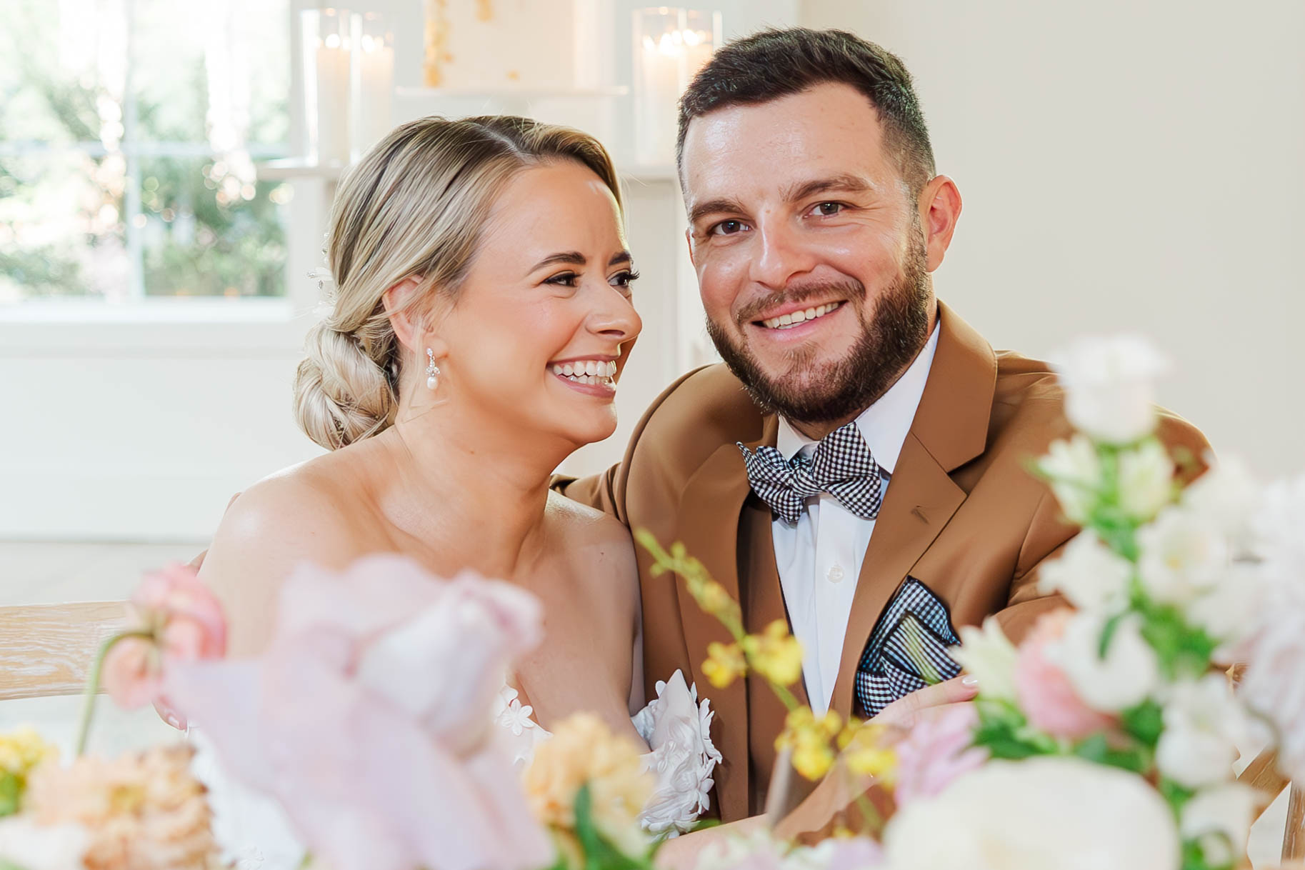 A smiling couple seated together, dressed elegantly for a wedding. The Bride, wearing a white floral gown and pearl earrings, looks joyfully at the Groom, who is dressed in a tan suit with a checkered bow tie. The foreground features soft, colorful flowers, and the background is softly lit with candles and natural light.