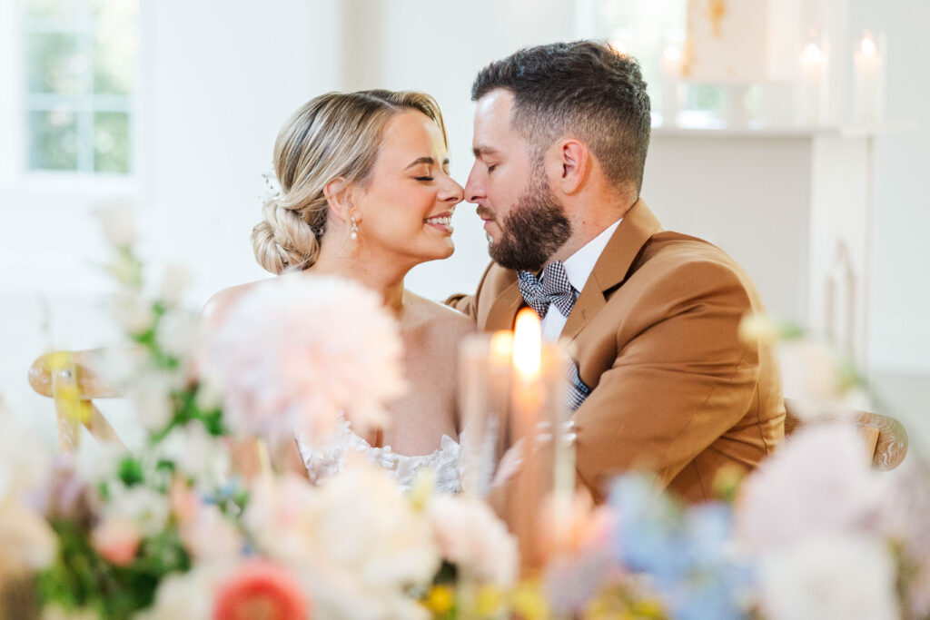 A bride and groom share an intimate moment, leaning in close with smiles, surrounded by soft, colorful floral arrangements and glowing candles. The bride wears a delicate white gown with her hair styled in a low bun, while the groom is dressed in a tan suit with a patterned bow tie. Captured by Virginia Wedding Photographer