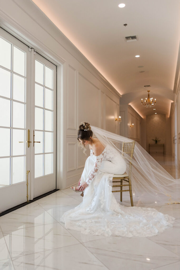 A bride sits on a gold chair in an elegant, light-filled hallway with white walls and marble floors, adjusting her shoe. She wears a stunning off-the-shoulder lace wedding gown with a long train and a flowing veil. The soft lighting and refined setting highlight the attention to detail captured by wedding photographers in Virginia Beach.