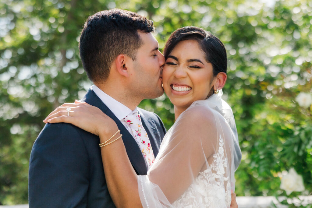 A groom kisses his smiling bride on the cheek during their wedding day. The bride, wearing a lace gown with sheer sleeves, beams with joy while holding the groom close. The groom is dressed in a dark suit with a floral tie. They are outdoors with lush greenery in the background.