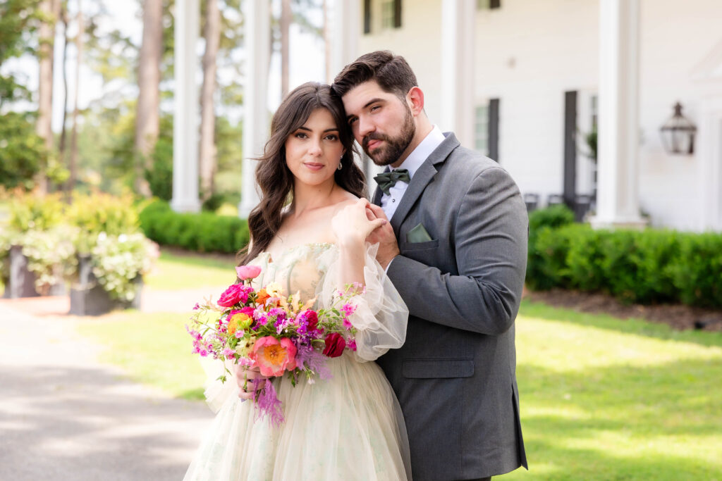 A couple poses outdoors in front of a white building with tall columns and lush greenery. The woman is wearing a light, flowing dress with sheer sleeves and holding a vibrant bouquet of pink, orange, and purple flowers. The man is dressed in a gray suit with a green bow tie and pocket square, gently holding the woman's shoulder. Both have a calm and elegant expression.