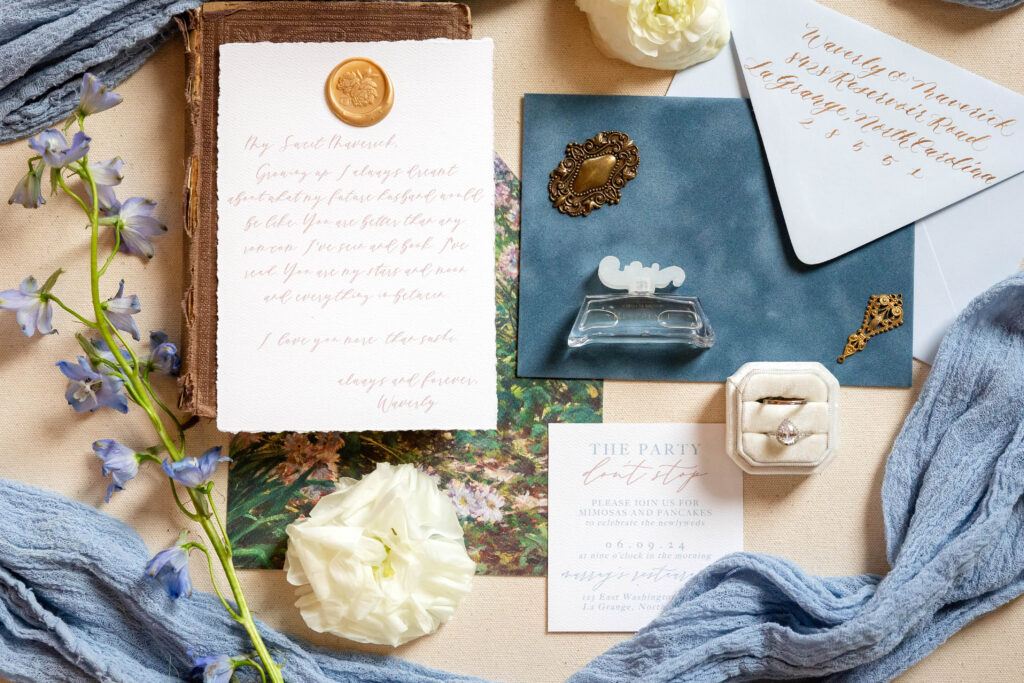 A flat-lay of elegant wedding details, including handwritten vows on textured paper sealed with a gold wax stamp, a blue envelope with gold calligraphy, a vintage gold brooch, and a white velvet ring box holding a pear-shaped diamond ring. Surrounding the arrangement are soft blue fabric, delicate purple flowers, and a white rose, creating a romantic and timeless aesthetic. This beautifully styled image highlights the artistry of Virginia Beach wedding photographers.