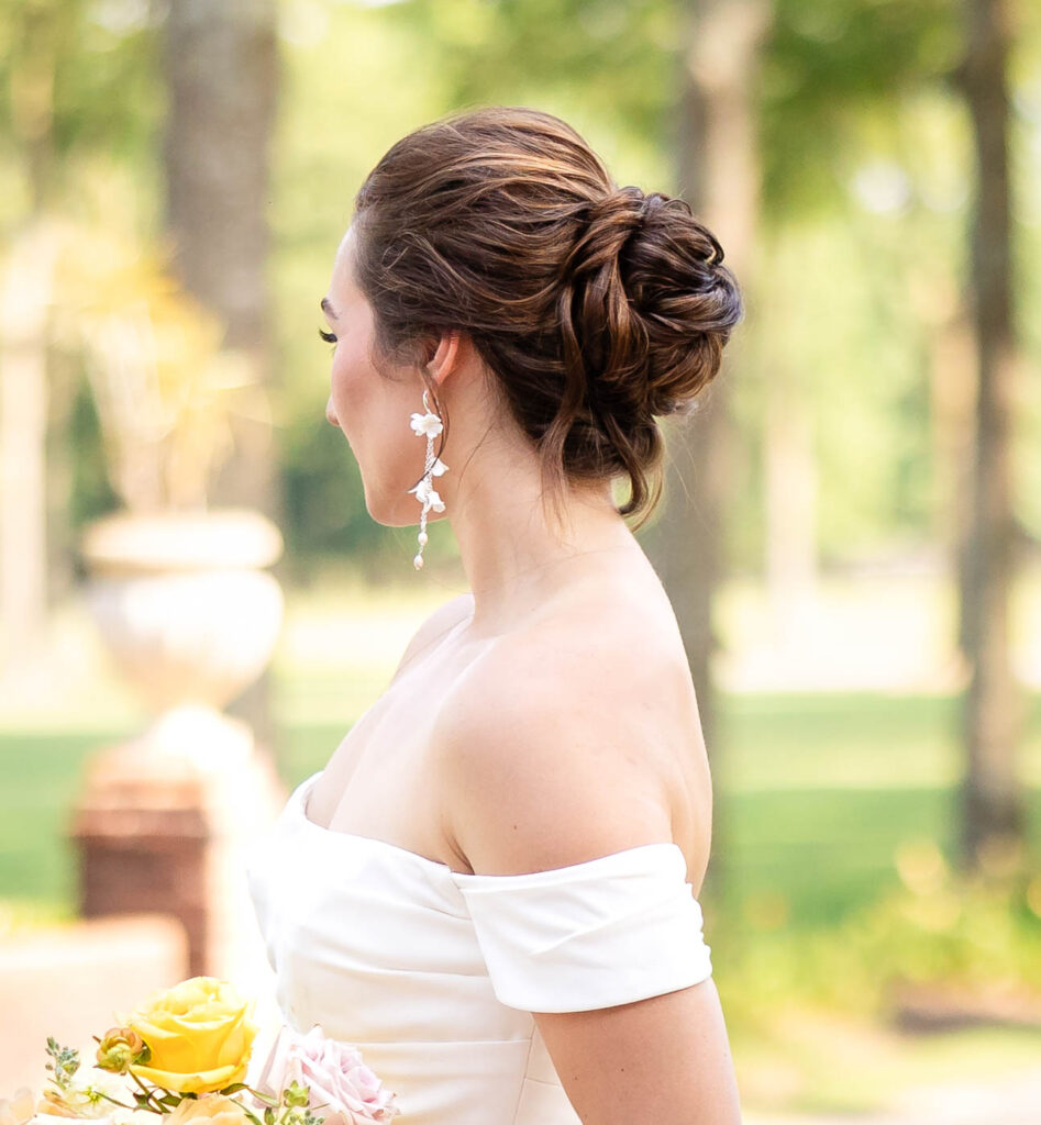 A bride stands outdoors in a sunlit garden, showcasing her elegant updo with intricate curls and delicate floral earrings. She wears an off-the-shoulder white wedding gown and holds a bouquet of yellow and blush roses. The soft greenery and blurred background create a romantic atmosphere, beautifully captured by a Virginia wedding photographer.