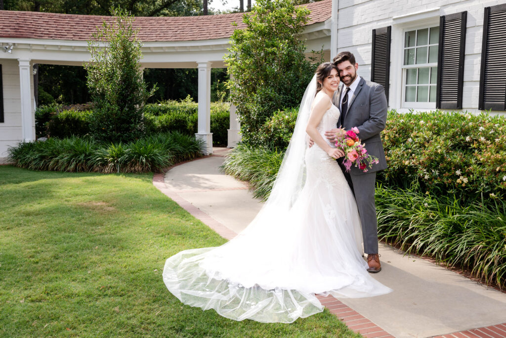 A bride and groom share an intimate moment in front of a charming white building with black shutters and lush greenery, captured on a sunny day. The bride wears a fitted lace wedding gown with a long train and veil, holding a vibrant bouquet of pink, orange, and red flowers. The groom is dressed in a gray suit with a black tie, gazing lovingly at the bride. This romantic outdoor scene highlights the work of a Virginia Beach wedding photographer.