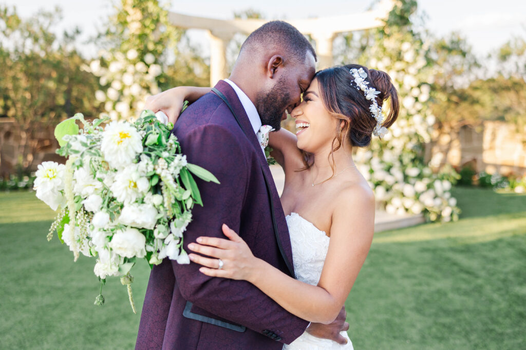 A joyful bride and groom embrace outdoors on their wedding day. The bride, wearing a strapless white lace gown and a floral hairpiece, smiles brightly as she holds a bouquet of white flowers. The groom, dressed in a deep purple suit, leans in with a loving expression. They are surrounded by greenery and floral decorations in a sunlit garden setting.