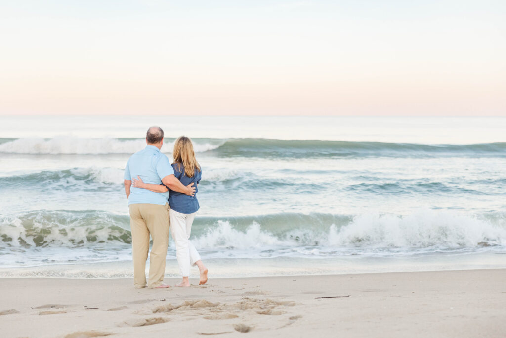 A couple stands barefoot on the sandy shore, embracing as they gaze at the gentle waves of the ocean under a pastel-colored sky. The serene beach setting captures a romantic and timeless moment, showcasing the artistry of Virginia Beach wedding photographers.