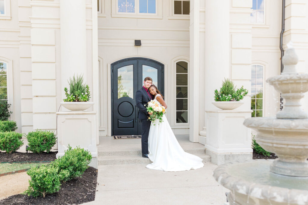 Bride and Groom standing in front of a white marbles estate with a double black door entrance.  Fountain in the foreground.