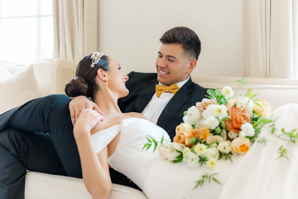 Bride and Groom relaxing on a couch after their wedding.  Bride holding her bouquet and they hold hands and look lovingly into each others eyes while smiling.  By Hampton Roads Wedding Photographers