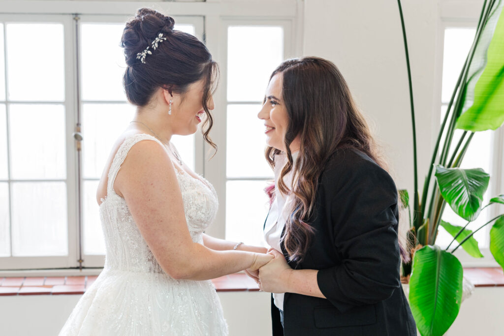 Bride holding hands with her sister in an emotional moment before the ceremony, standing in a bright room with large windows and lush greenery at the Virginia Cavalier