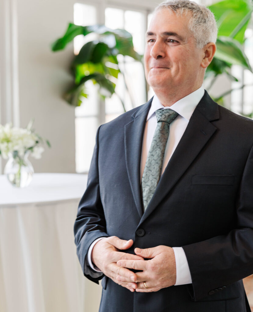 Father of the bride smiling proudly, standing in a bright room with soft natural light and elegant floral decor.  This is in the pool and terrace at the Virginia Cavalier.