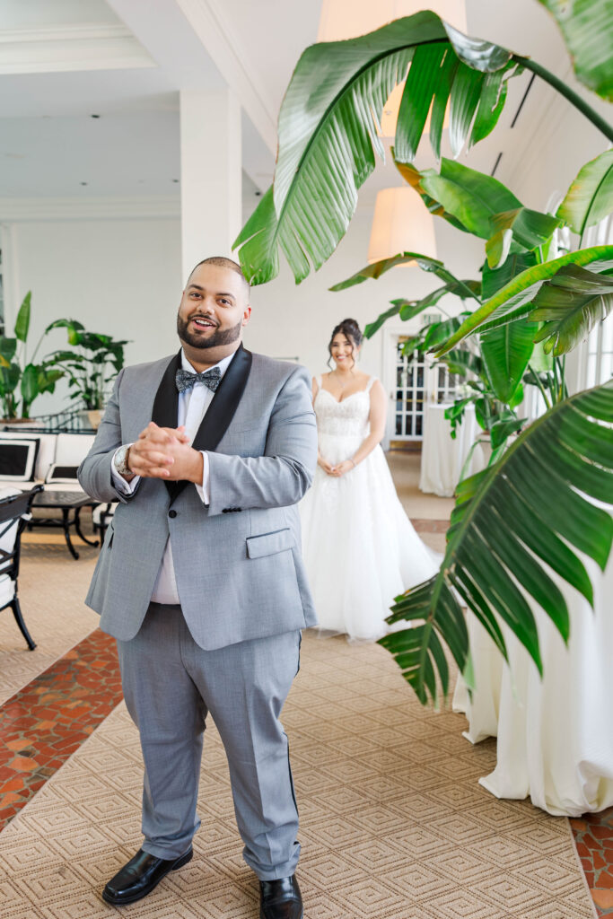 Groom standing in anticipation during the first look, with the bride approaching from behind in a beautifully lit room with tropical plants.  Room is at the Virginia Cavalier.