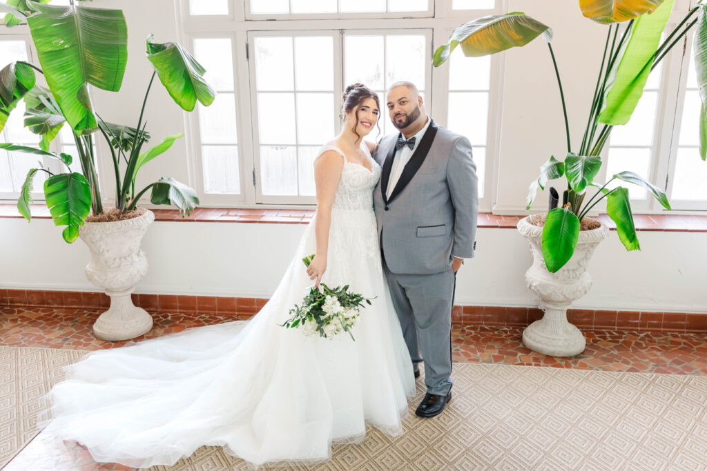 Bride and groom posing in a bright, airy room with tropical plants and large windows, showcasing the timeless elegance of The Virginia Cavalier.