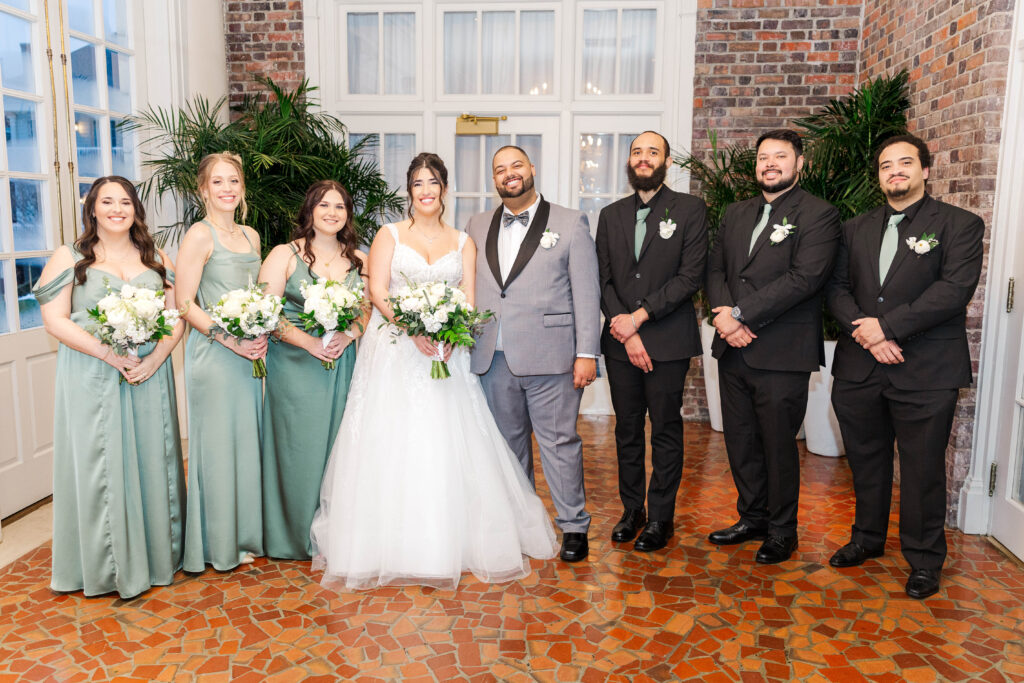 Bride and groom posing with their bridal party, bridesmaids in sage green dresses and groomsmen in black suits, in the elegant interior of The Virginia Cavalier.