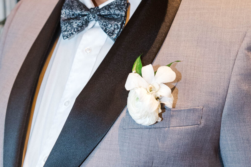 Close-up of the groom’s boutonniere, a white flower pinned to a light gray suit jacket, paired with a patterned bow tie at The Virginia Cavalier.