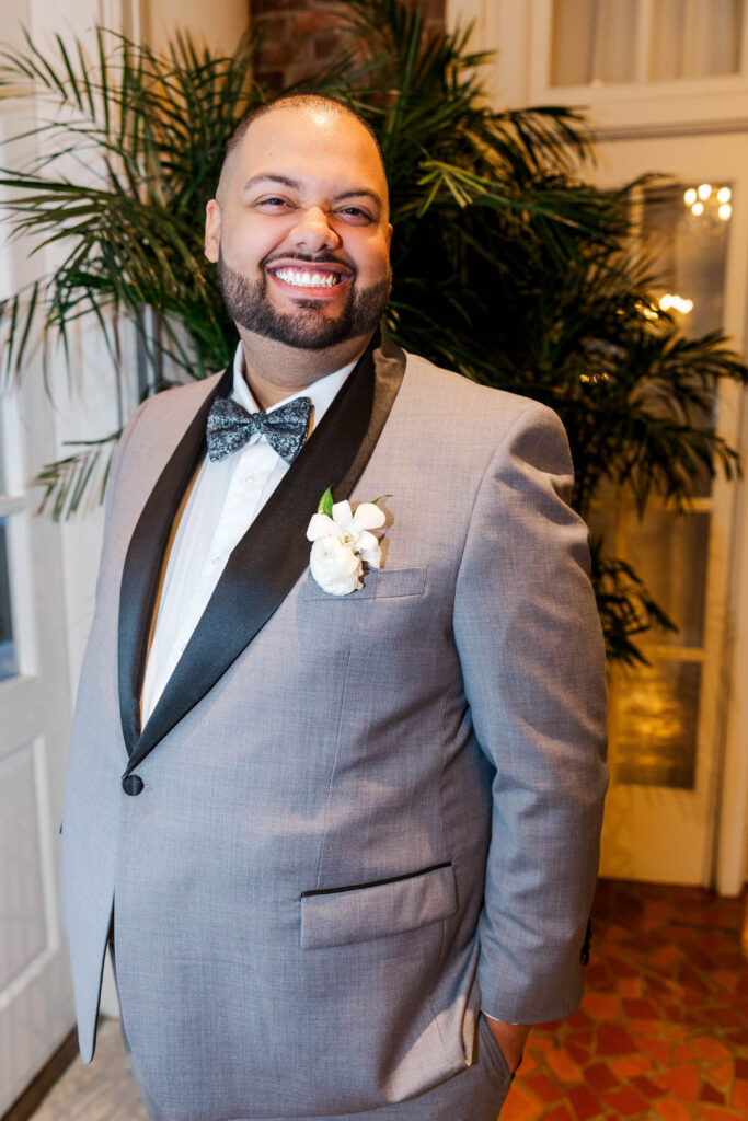 Groom smiling brightly in a light gray suit with a black lapel, standing in front of lush greenery and elegant decor at The Virginia Cavalier.